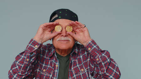 Happy one man winner showing golden bitcoins. Career wealth, cryptocurrency investment, mining future technology. Elderly grandfather fooling making faces. Senior businessman on gray studio backgroundの写真素材