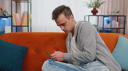 Smiling Caucasian young adult man sitting on couch using credit bank card and smartphone while...