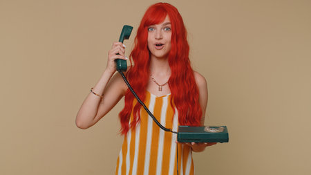 Hey you, call me back. Cheerful one ginger woman secretary in tank top talking on wired vintage telephone of 80s, says hey you call me back. Young redhead girl posing isolated on beige wall backgroundの写真素材