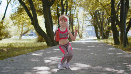 Athletic sport runner child girl training fitness exercise listening Bluetooth music speaker dancing to camera having fun. Workout cardio in park. Toddler kid jogger enjoy funny dance. Active childrenの写真素材