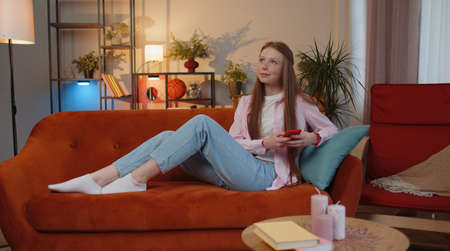 Young teen child girl lying on sofa uses mobile phone smile at modern home apartment. Ginger kid texting share messages content on smartphone social media applications online, watching relax movieの写真素材