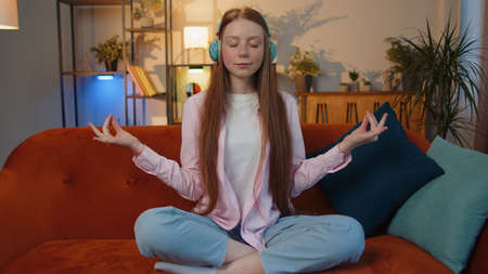 Keep calm down, relax, inner balance. Young redhead child girl breathes deeply with mudra gesture eyes closed meditating with concentrated thoughts, peaceful mind sit at home in living room on couchの写真素材