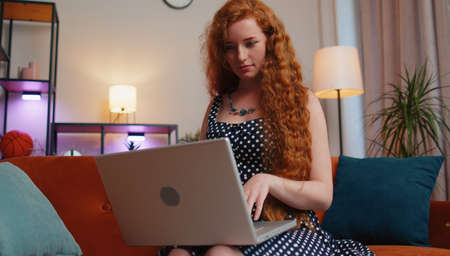 Redhead young woman in polka dots dress freelancer in home living room sits on couch, opens laptop start working. Ginger girl works on notebook, sends messages, makes online purchases, watching moviesの写真素材