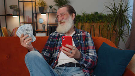 Planning family budget. Smiling senior old grandfather counting money cash, use smartphone calculate...