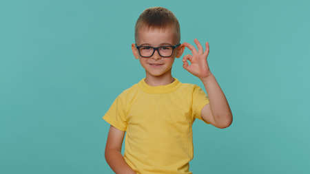 Ok. Little toddler children boy looking approvingly at camera showing agree gesture, like sign positive, approve something good. Young preschool kid in glasses isolated alone on blue studio backgroundの写真素材