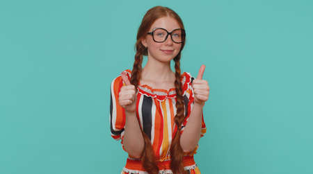 Great nice job. Cheerful positive happy girl in dress showing thumbs up and nodding in approval, successful good work. Young stylish ginger teenager child on blue background. People sincere emotionsの写真素材