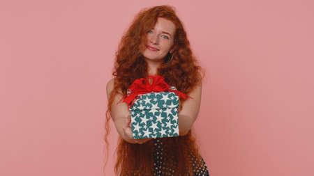 Smiling redhead young woman presenting birthday gift box stretches out hands offer wrapped present...