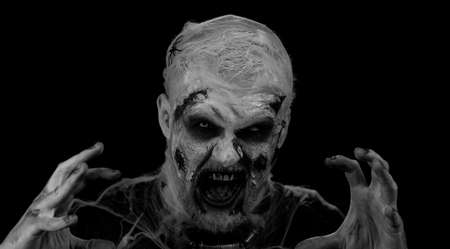 Creepy man with Halloween zombie bloody wounded makeup, trying to scare, face expressions. Horror theme. Sinister undead guy isolated on studio black background. Voodoo rituals. Fashion body artの写真素材