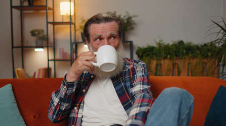 Attractive smiling senior old grandfather man drinking a cup of warm coffee or herbal tea sitting at home living room couch in the morning. Elderly retired grandpa pensioner enjoying comfortable relaxの写真素材