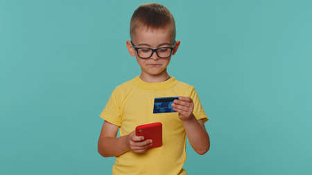 Little toddler children boy in yellow t-shirt using credit bank card and smartphone while transferring money, purchases online shopping. Young preschool kid isolated alone on blue studio backgroundの写真素材