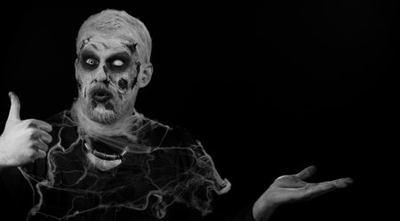 Zombie man with makeup with fake wounds scars showing thumbs up and pointing empty place,...