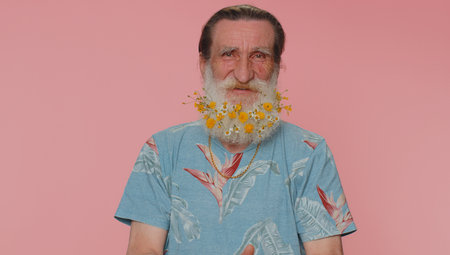 Cheerful lovely elderly man with beard with flowers in blue t-shirt smiling and looking at camera. Senior pensioner model grandfather indoor studio shot isolated on pink background. Male nature beautyの写真素材