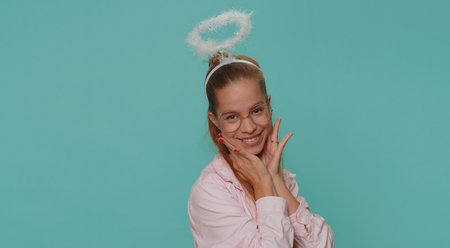 Portrait of smiling shy angelic pretty teenager young girl with angel halo nimb over head flirting, looking at camera, positive emotions, celebrating holiday. Student child kid on blue backgroundの写真素材
