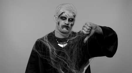 Sinister man in carnival costume of Halloween crazy zombie with bloody wounded scars face showing thumbs down sign gesture, expressing discontent, disapproval, dissatisfied, dislike. Cosplay undeadの写真素材