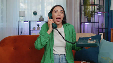 Crazy adult girl in casual shirt talking on wired vintage old-fashioned retro telephone of 80s, fooling, making silly faces. Young woman sitting on orange couch at home living room making conversationの写真素材