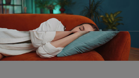 Tired caucasian adult girl lying down in bed taking a rest at home. Carefree young woman napping, falling asleep on comfortable orange sofa with pillows. Closed her eyes enjoy daytime nap aloneの写真素材