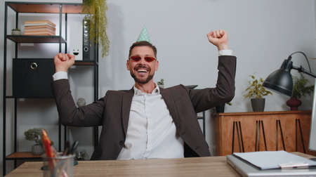 Businessman wears sunglasses, festive cap, listening music at workplace dancing victory dance, weekends. Rejoicing developing project. Celebrating success. Lottery jackpot. Motivated people in officeの写真素材