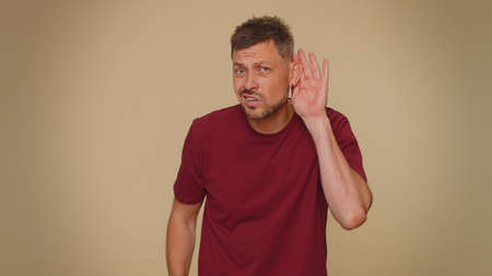 I cant hear you. What. Young man trying hear you, looking confused and frowning, keeping arm near ear for louder voice, asking to repeat, to hear information, deafness. One guy on beige backgroundの写真素材