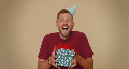 Cheerful bearded handsome young man receiving gift box and smiling joyfully, looking amazed satisfied with nice present, unexpected birthday surprise. Stylish male guy on beige studio backgroundの写真素材