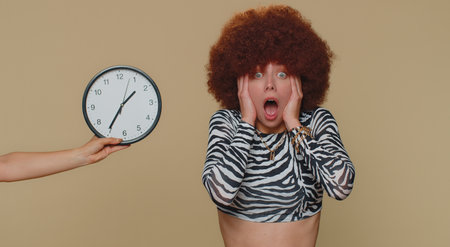 Young woman in brown lush wig with anxiety checking time on clock, running late to work, being in...