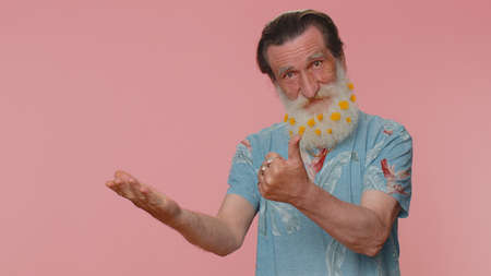 Handsome senior man showing thumbs up and pointing empty place, advertising area for commercial text, copy space for goods promotion. Elderly grandfather. Studio shot indoors on pink wall backgroundの写真素材