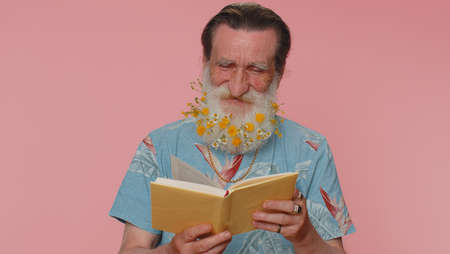 Senior man with flowers in gray-haired beard reading funny interesting fairytale story book, leisure hobby, knowledge wisdom, education, learning, study, wow. Elderly grandfather on pink backgroundの写真素材