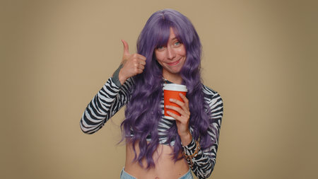 Positive pretty young woman drinking coffee from cup. Adult stylish girl enjoying hot beverage in papercup, holding espresso in hand on beige studio background. Energy and relax with refreshment drinkの写真素材