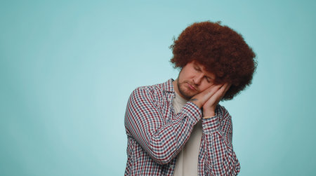 Tired handsome bearded man with lush hair coiffure yawning, sleepy inattentive feeling somnolent lazy bored gaping suffering from lack of sleep. Young guy boy isolated alone on blue studio backgroundの写真素材