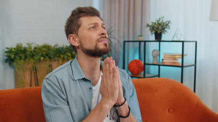 Please, God help. Man praying sincerely with folded arms, looking upward and making wish, asking with hopeful imploring expression, begging apology. Young guy sitting on couch at home living roomの写真素材