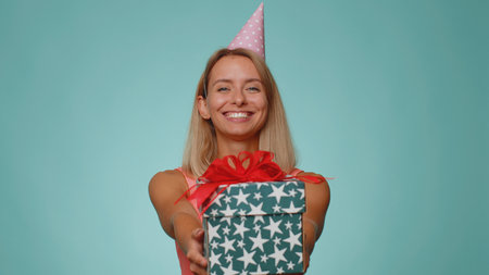 Positive smiling blonde woman presenting birthday gift box stretches out hands, offer wrapped present career bonus, celebrating party. Young adult girl isolated alone on blue studio wall backgroundの写真素材
