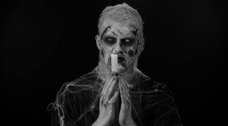 Frightening man with Halloween zombie bloody wounded makeup spells conjures over a candle. Horror theme. Sinister undead guy isolated on studio black background. Voodoo rituals. Fashion body artの写真素材
