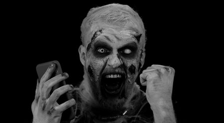 Happy excited sinister man in carnival costume of Halloween crazy zombie looking smartphone display sincerely rejoicing win, receiving message with good news, success luck. Cosplay wounded undeadの写真素材
