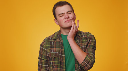 Teenager young man touching sore cheek suffering from toothache cavities or gingivitis waiting for dentist appointment, gums disease. Adult boy indoors studio shot isolated alone on yellow backgroundの写真素材