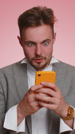 Worried amazed bearded business man enthusiastically playing racing or shooter video games on smartphone. Young guy using mobile phone gadget celebrate lottery jackpot, good news, goal. Vertical viewの写真素材