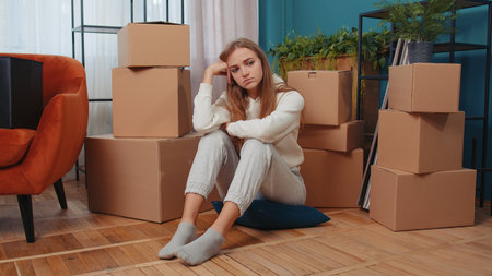 Upset depressed young woman sitting on floor near cardboard boxes with stuff. Sad girl tired of long relocation, bankruptcy, desperate due lender repossess home, bank house loan debt, divorce conceptの写真素材