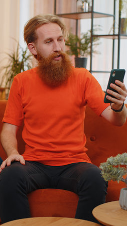 Portrait of caucasian bearded man making smartphone call conversation with friends. Happy excited young guy at home in room on orange couch enjoying mobile phone loudspeaker talking. Vertical viewの写真素材