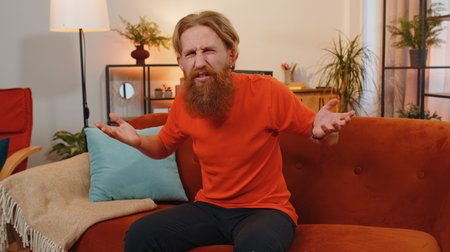 What. Why. Bearded ginger man raising hands in indignant expression, asking reason of failure demonstrating disbelief irritation by troubles at home apartment. Young guy in room sitting on orange sofaの写真素材