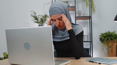 Tired muslim business woman in hijab use laptop suffering from headache problem tension, migraine, stress. Girl freelancer works on notebook, sends messages, online purchases at home office workspaceの写真素材