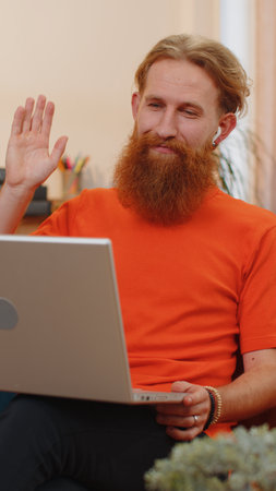 Portrait of bearded redhead man sitting on couch, looking at camera, making video webcam conference call with friends or family enjoying pleasant conversation. Young guy laughing, waving hello at homeの写真素材