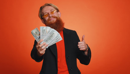 Rich pleased boss bearded man waving money dollar cash banknotes bills like a fan, success business career, lottery winner, big income, wealth. Young guy boy isolated alone on orange studio backgroundの写真素材