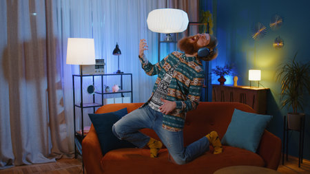 Happy hippie rocker young man in wireless headphones relaxing at home dancing on couch listening...