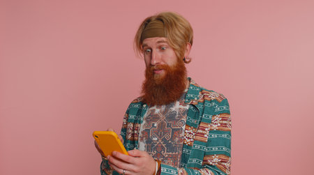 Happy excited joyful hippie man in pattern shirt use mobile cell phone typing browsing shouting say wow yes found out great big win, good news, lottery goal achievemen, celebrating success, winningの写真素材