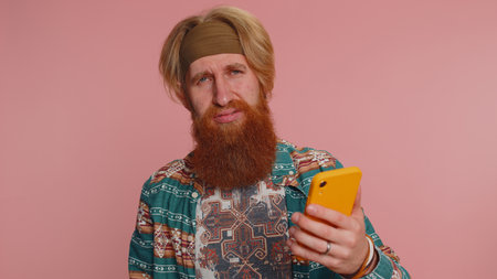 Bearded hippie man in pattern shirt use mobile cell phone typing browsing, loses becoming surprised sudden lottery results, bad fortune, loss, fail. Young redhead guy boy isolated on pink backgroundの写真素材