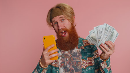 Happy hippie tourist man looking smartphone display sincerely rejoicing win, receiving money dollar...