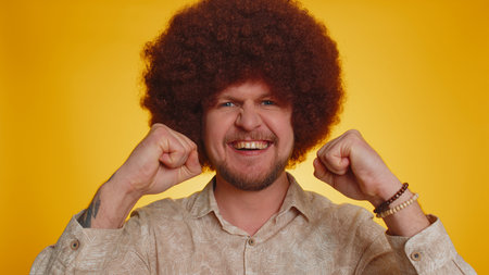 Happy man with lush afro hairstyle coiffure shouting, raising fists in gesture I did it, celebrating success, winning, birthday, lottery goal achievemen. Hipster guy boy on yellow studio backgroundの写真素材