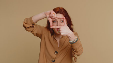 Redhead woman photographer gesturing picture frame with hands, looks through fingers and focusing on...