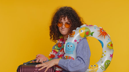 Happy curly haired woman in sunglasses, hawaiian lei walking along with luggage bag, swimming ring....