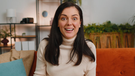 Oh my God, Wow. Surprised woman with brunette hair at home couch, shocked by sudden victory triumph,...