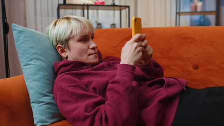 Young woman with short blonde hair lying on sofa uses mobile phone smiles at night evening home....