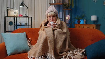 Sick young woman wrapped in plaid sitting, shivering from cold on sofa drinking hot tea in unheated apartment without heating due debt. Unhealthy girl feeling discomfort try to warm up at evening homeの写真素材